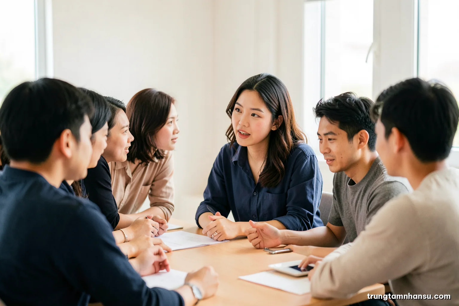 Small Vietnamese product team in a bright meeting room, one young woman speaking while colleagues lean in attentively, soft daylight, warm neutral palette, eye-level wide shot, faces partly turned away