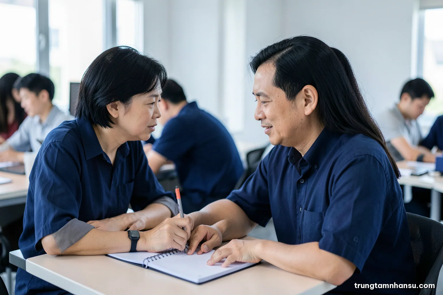 Two Vietnamese colleagues sitting side by side at a shared workstation, the senior one pointing at a blank notebook while explaining, side profile, cool daylight, navy and soft grey palette