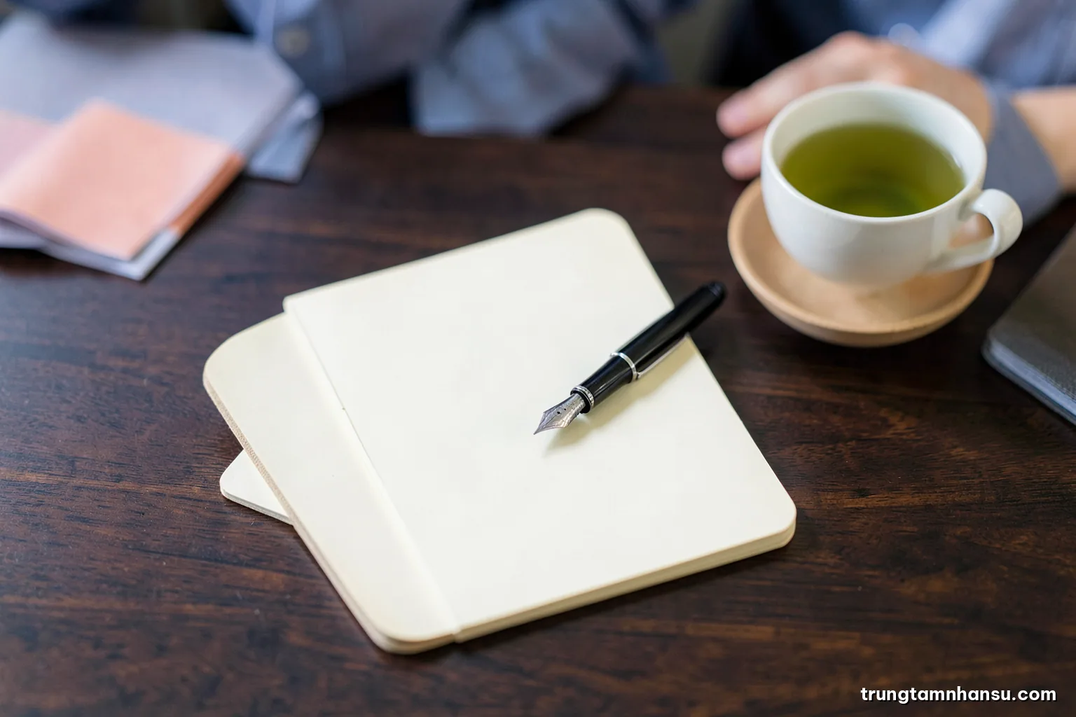 Top-down macro of a fountain pen resting on a blank cream notepad beside a cup of green tea on a dark walnut desk, soft diffused light, minimal composition