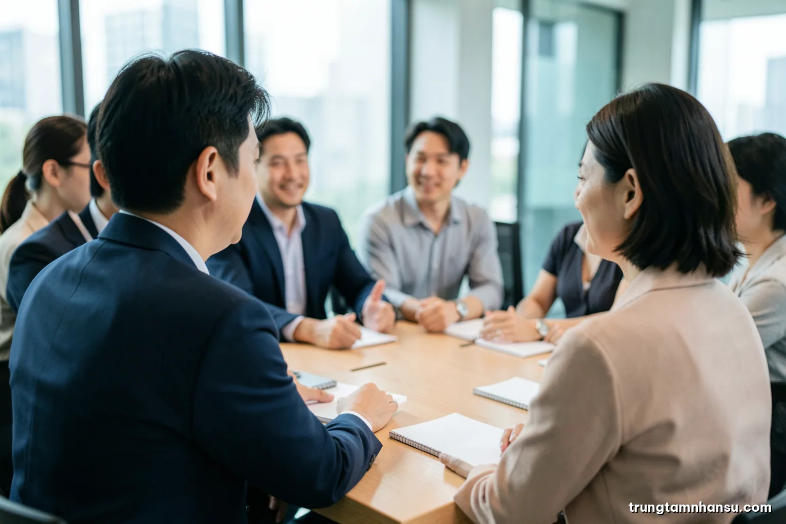 Over-the-shoulder view of a Vietnamese manager and employee talking across a light wooden table in a quiet glass meeting room, blank notebooks, soft morning daylight, muted teal palette