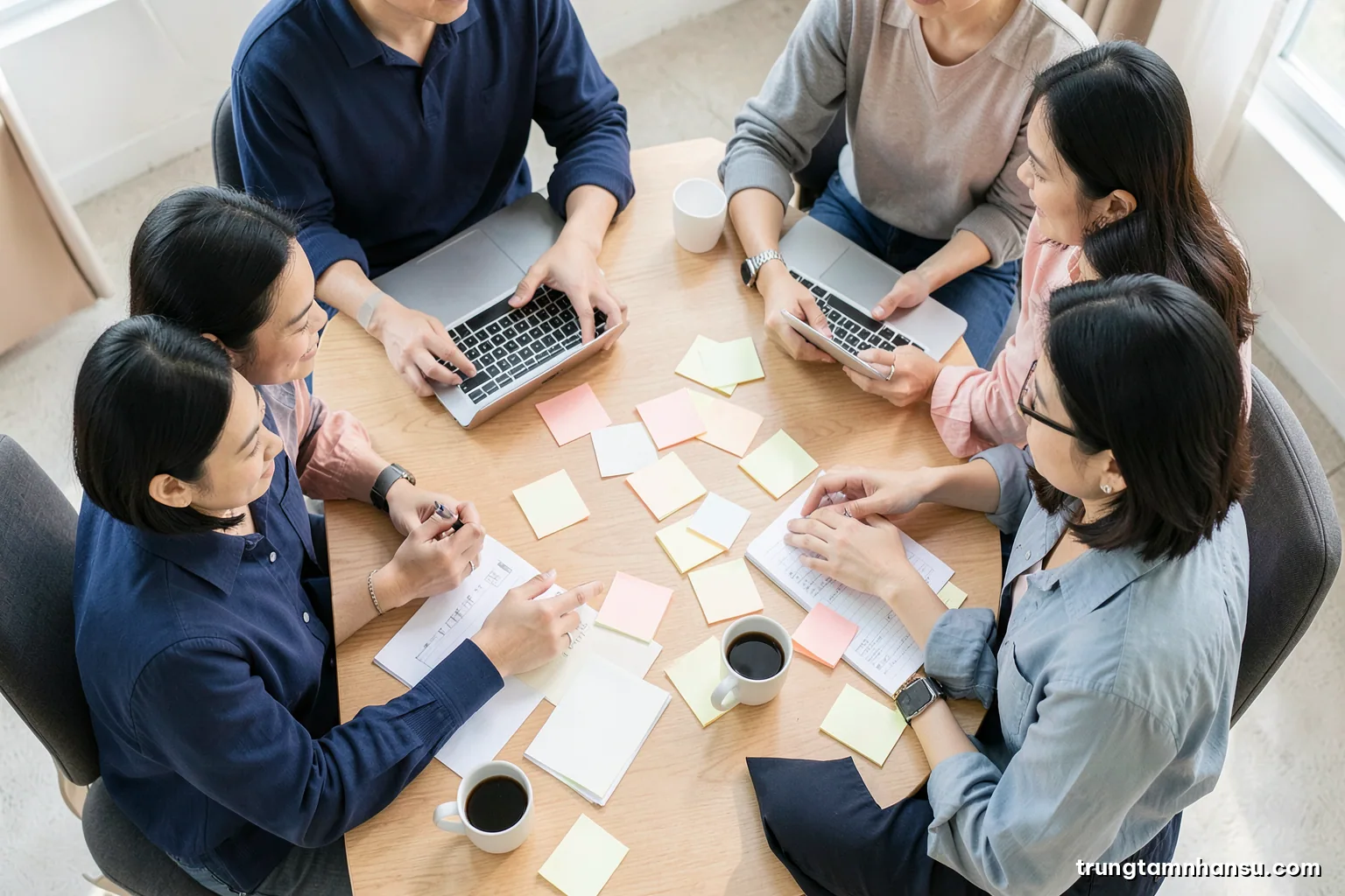 Overhead view of four Vietnamese professionals around a wooden table scattered with blank sticky notes, coffee cups and closed laptops, natural daylight, soft pastel palette