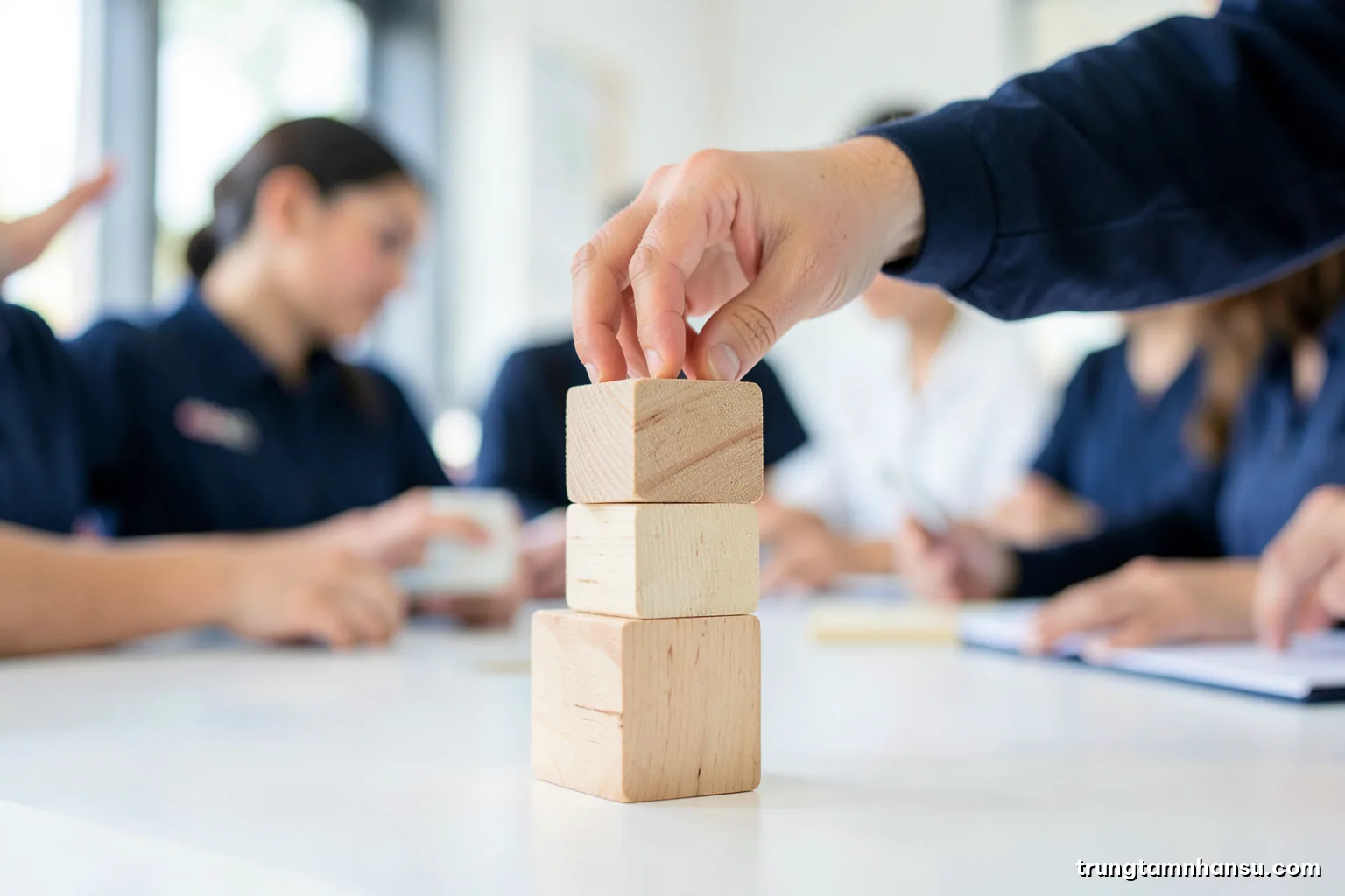 Low angle shot of three stacked wooden blocks of different heights on a white desk, a hand placing the top block, clean studio light, navy and pale sand palette