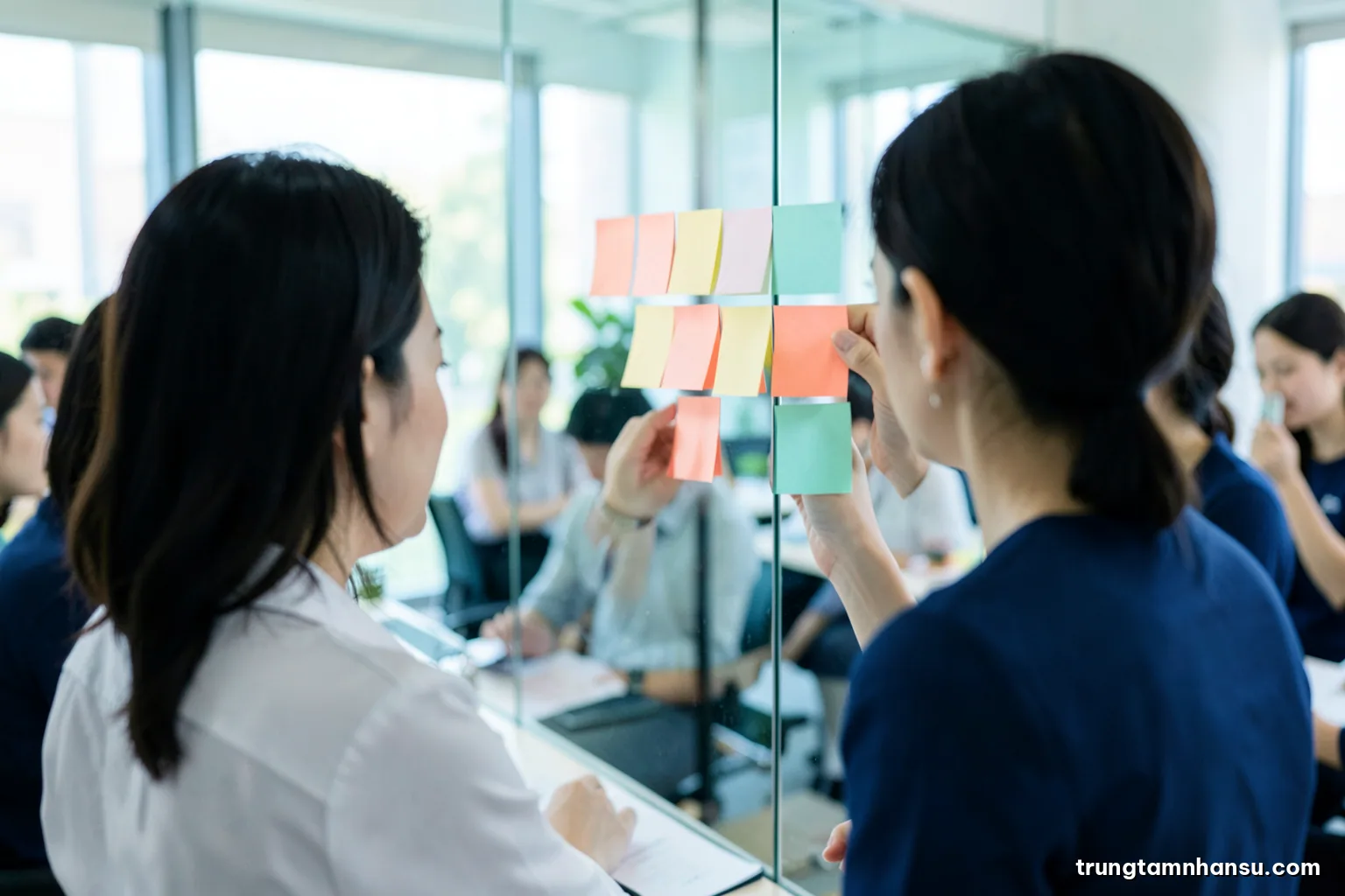 Over-the-shoulder view of a Vietnamese analyst arranging colored sticky notes into clusters on a glass wall, notes blank, bright office, teal and coral palette