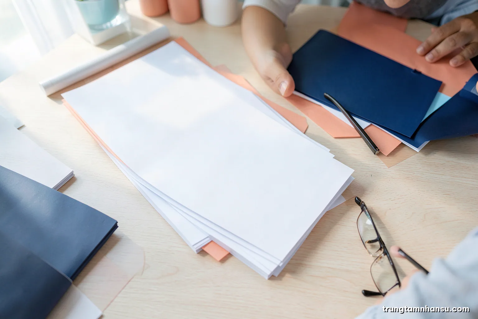 Top-down view of neatly stacked blank paper folders and a pair of reading glasses on a light birch desk, crisp morning light from the left, white and pale blue palette
