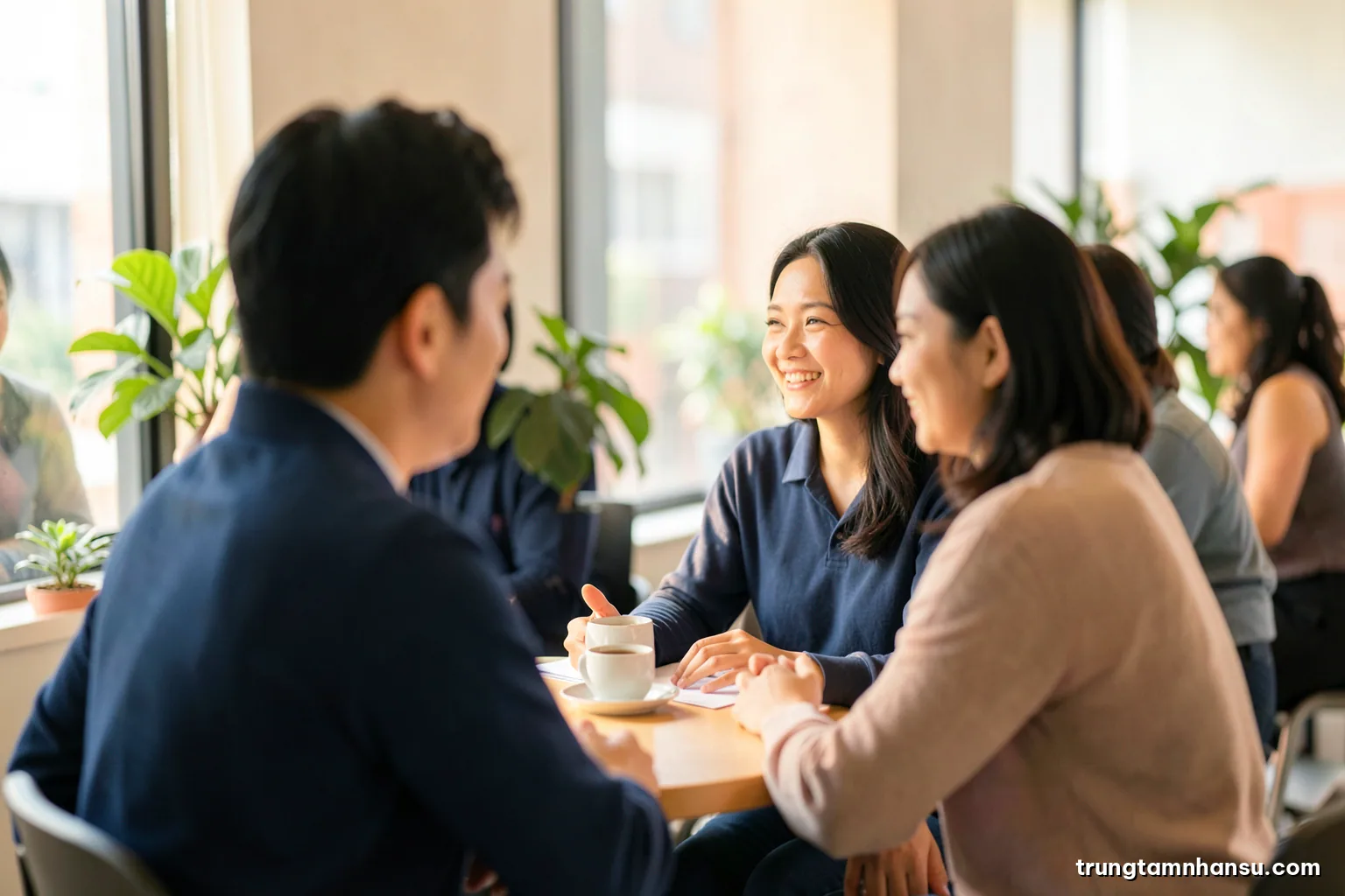 Two Vietnamese colleagues talking over coffee at a small round table by a large window, shot over the shoulder, faces softly out of focus, warm afternoon light, green plants