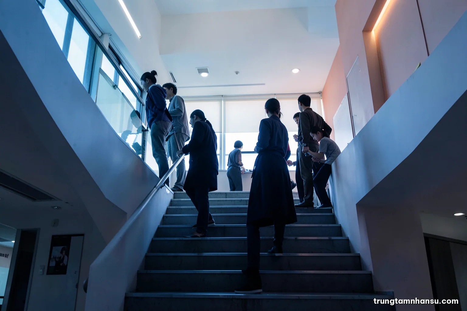 Low angle view of a staircase in a modern Vietnamese office atrium, several silhouetted figures standing on different steps, cool blue light and long shadows, minimal composition